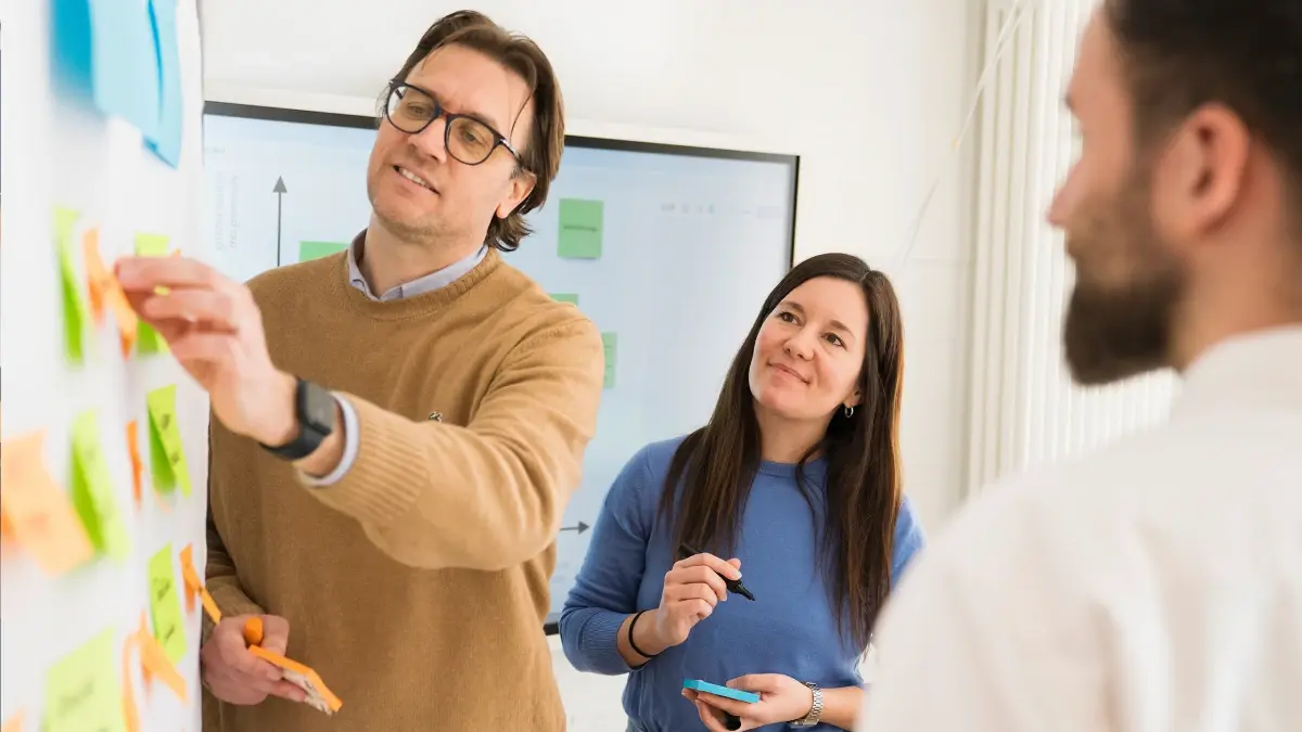 Drei Personen arbeiten in einem Büro zusammen und stehen vor einem Whiteboard, das mit bunten Haftnotizen bedeckt ist. Ein Mann bringt eine Notiz auf der Tafel an, während eine Frau und eine dritte Person engagiert lächelnd zuschauen.