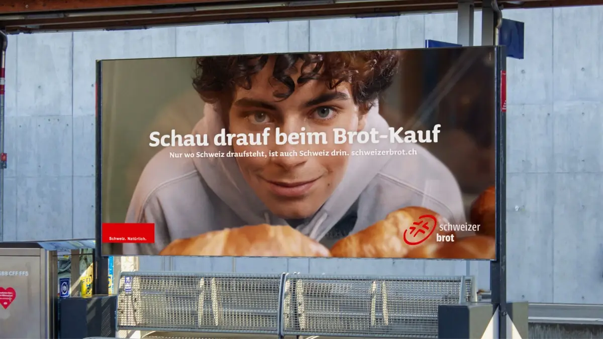 Ein Plakat zeigt einen jungen Mann, der frische Brötchen mit der Aufschrift "Schau drauf beim Brot-Kauf" und dem Logo von Schweizer Brot anlächelt und damit für Schweizer Brot als Teil einer nationalen Kampagne an einem Bahnhof wirbt.