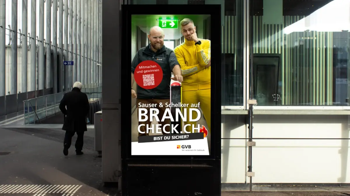 Eine digitale Plakatwand an einer Bushaltestelle zeigt eine Anzeige mit zwei Männern - einem in einem schwarzen Hemd und einem in einem gelben Overall -, die neben einem roten Notfallknopf stehen. Die Anzeige wirbt für "Brandcheck.ch" zur Sensibilisierung für den Brandschutz.