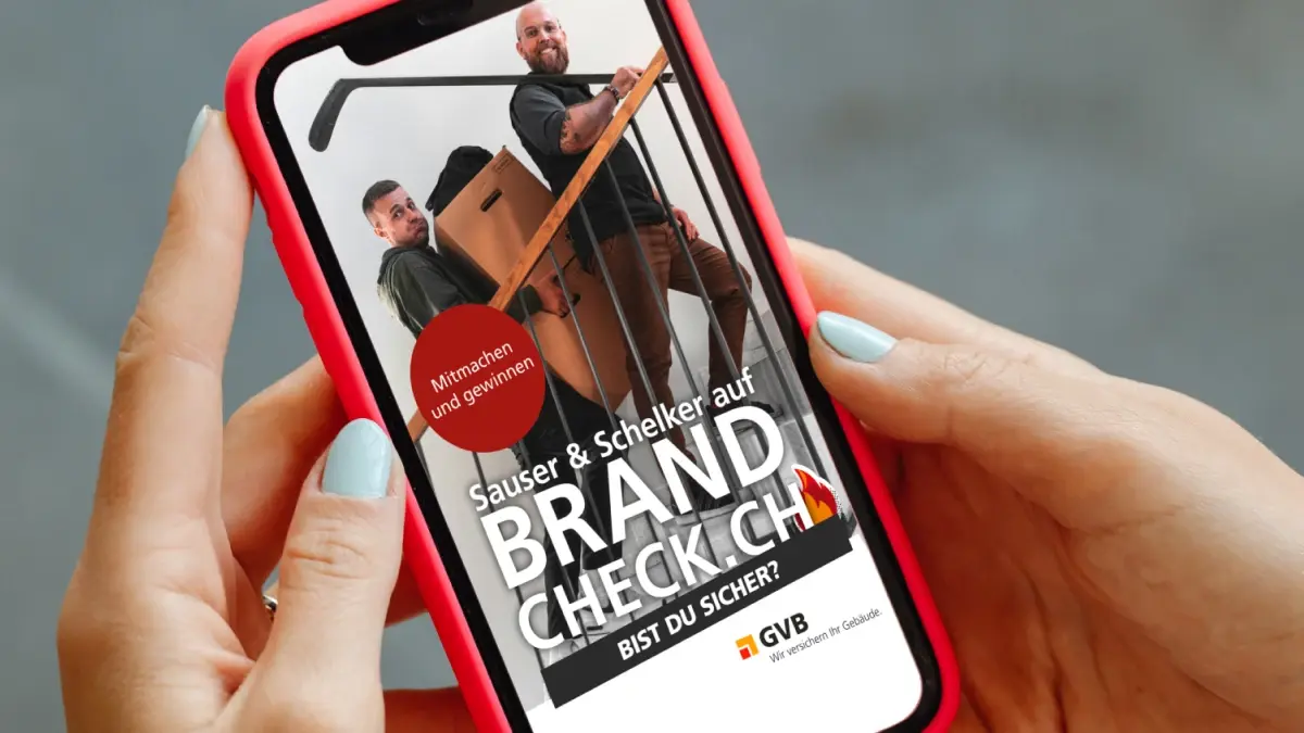 Eine Person hält ein Smartphone in der Hand, auf dem eine Anzeige mit zwei Männern zu sehen ist, die eine Couch nach oben tragen. Der Text lautet: "Sauser & Schelker auf Brand Check. Bist du sicher? Mitmachen und gewinnen! GVB.".