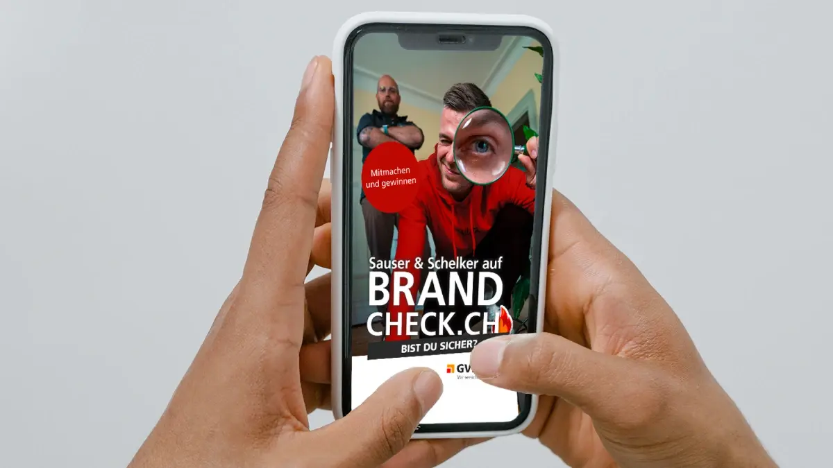 Eine Person hält ein Smartphone in der Hand, das eine Anzeige mit zwei Männern zeigt, von denen einer durch ein Vergrößerungsglas schaut. Der Text auf dem Bildschirm lautet: "Sauser & Schelker auf BRANDCHECK.CH Bist du sicher?" mit einem roten Kreis und Logos am unteren Rand.